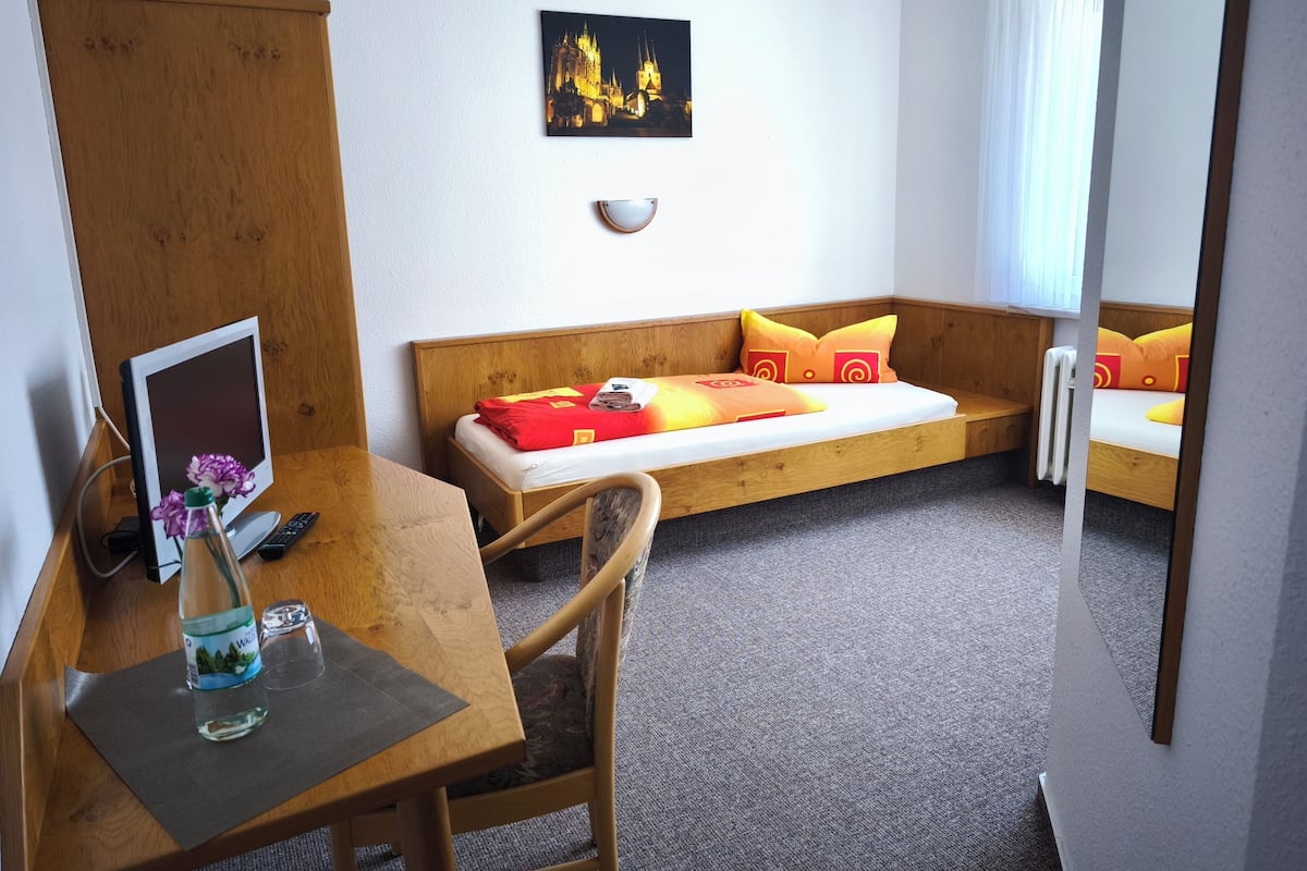 Ein Hotelzimmer mit einem Holztisch, einem Bett mit orangefarbener und gelber Bettwäsche, einem Gemälde einer Burg und einem Fenster mit weißen Vorhängen.