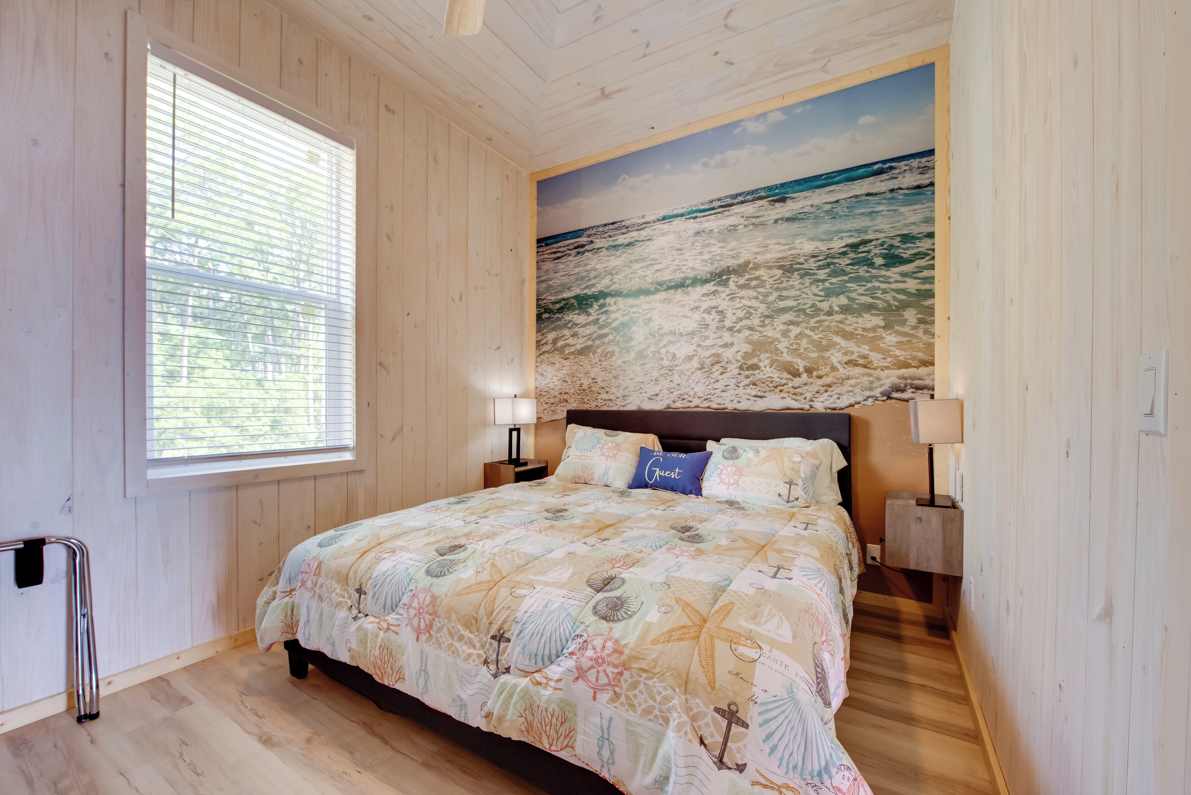 Un dormitorio con piso de madera, una cama con cubrecama de temática náutica, una ventana con persianas y un mural marino en la pared.