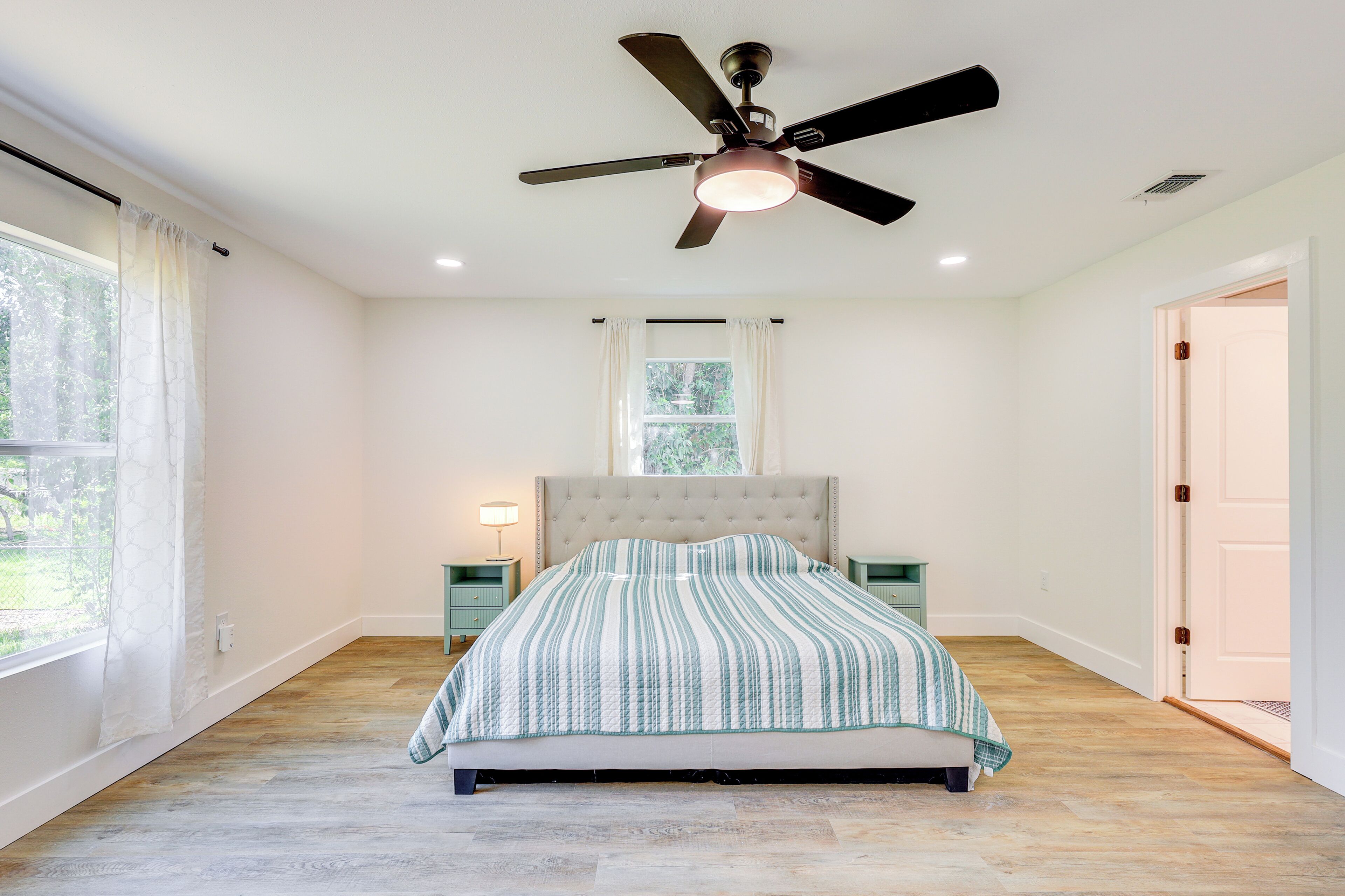 A bedroom with a bed, a ceiling fan, a window with curtains, and a nightstand with a lamp.