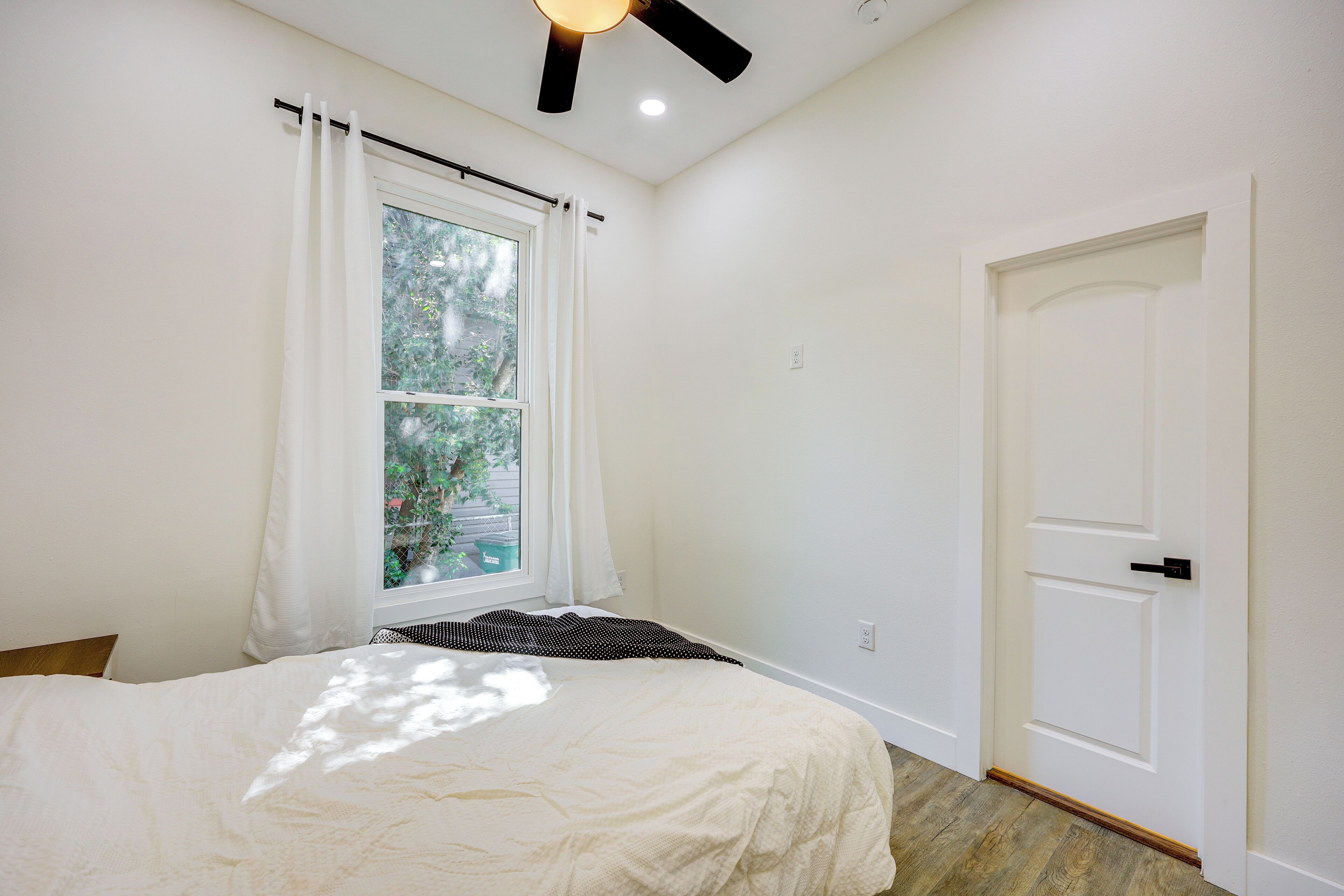 A bedroom with a bed, a window with curtains, a ceiling fan, and a door.
