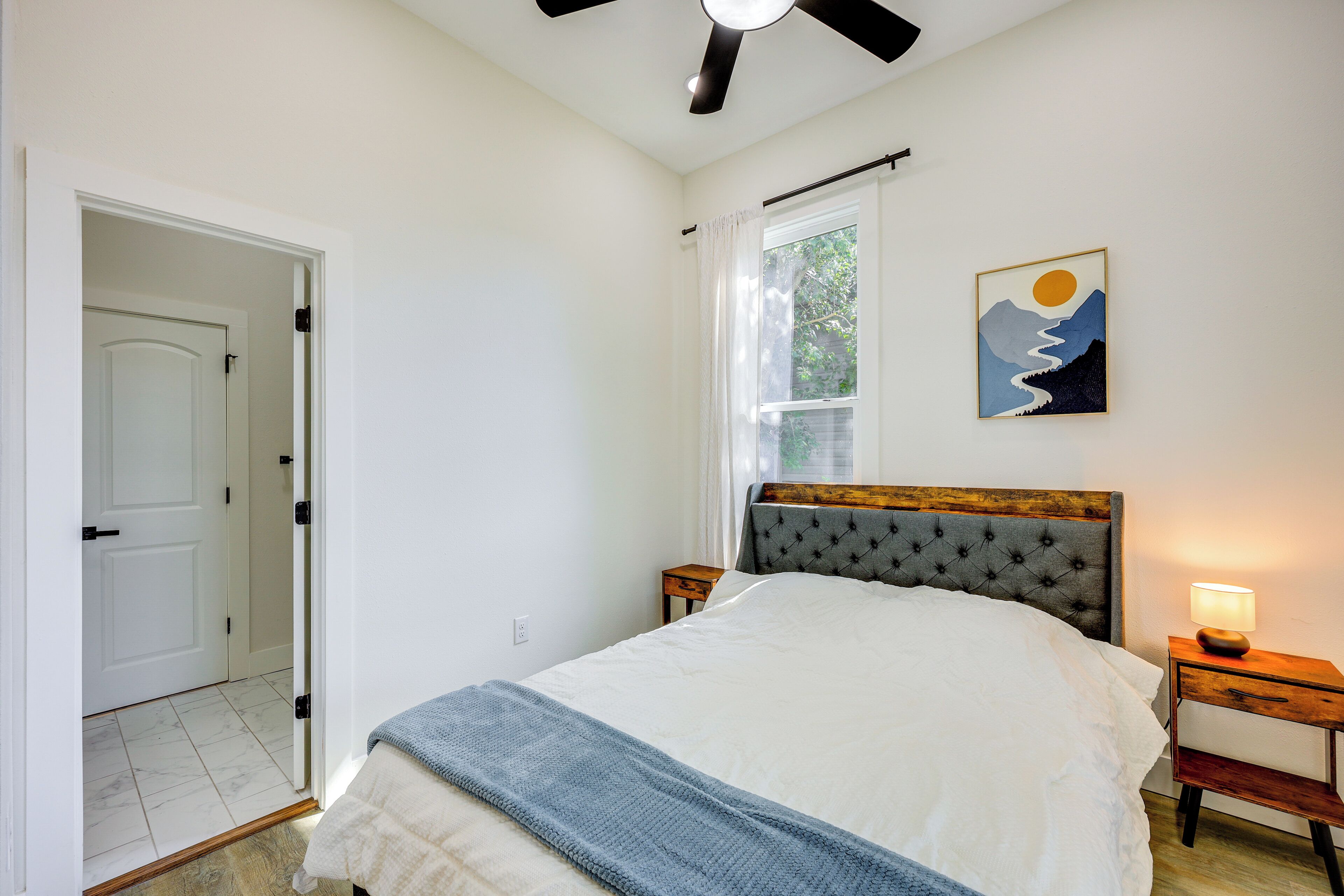 A bedroom with a bed, a nightstand, a ceiling fan, a window with curtains, and a wall-mounted artwork.