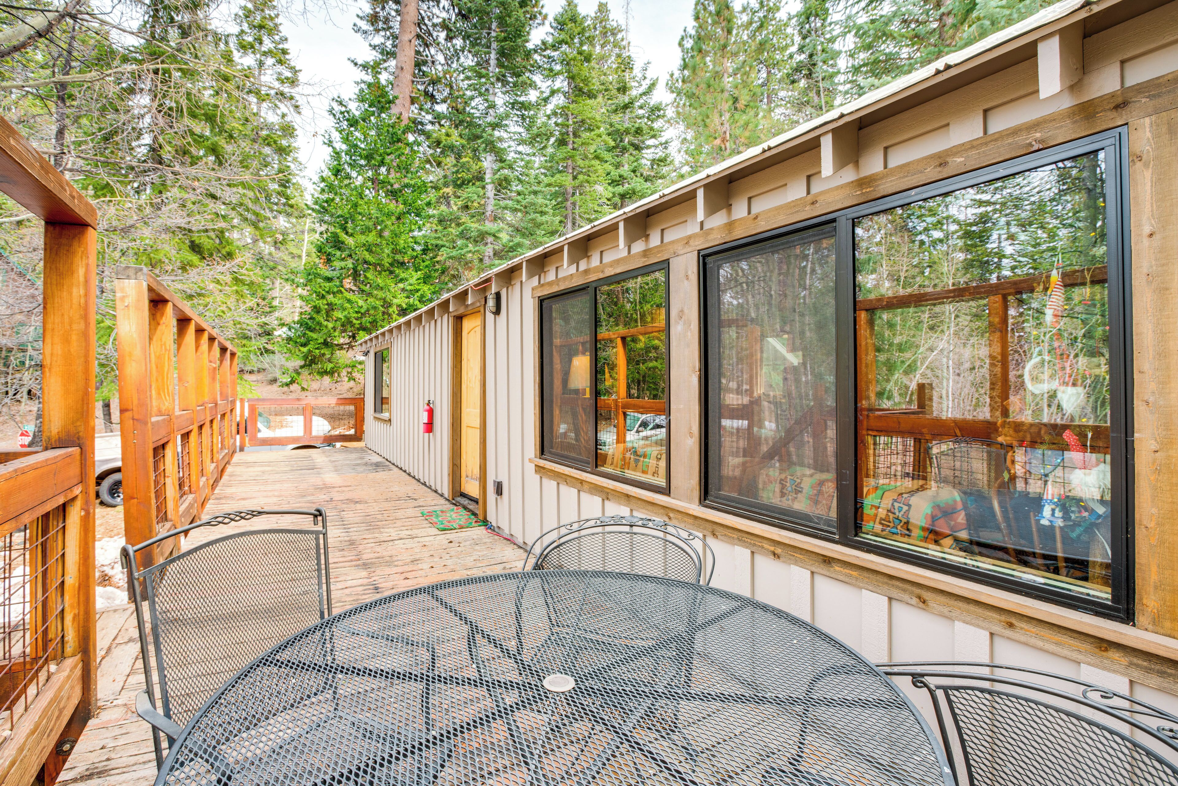 A wooden deck with metal tables and chairs, large windows, and a view of a forest.