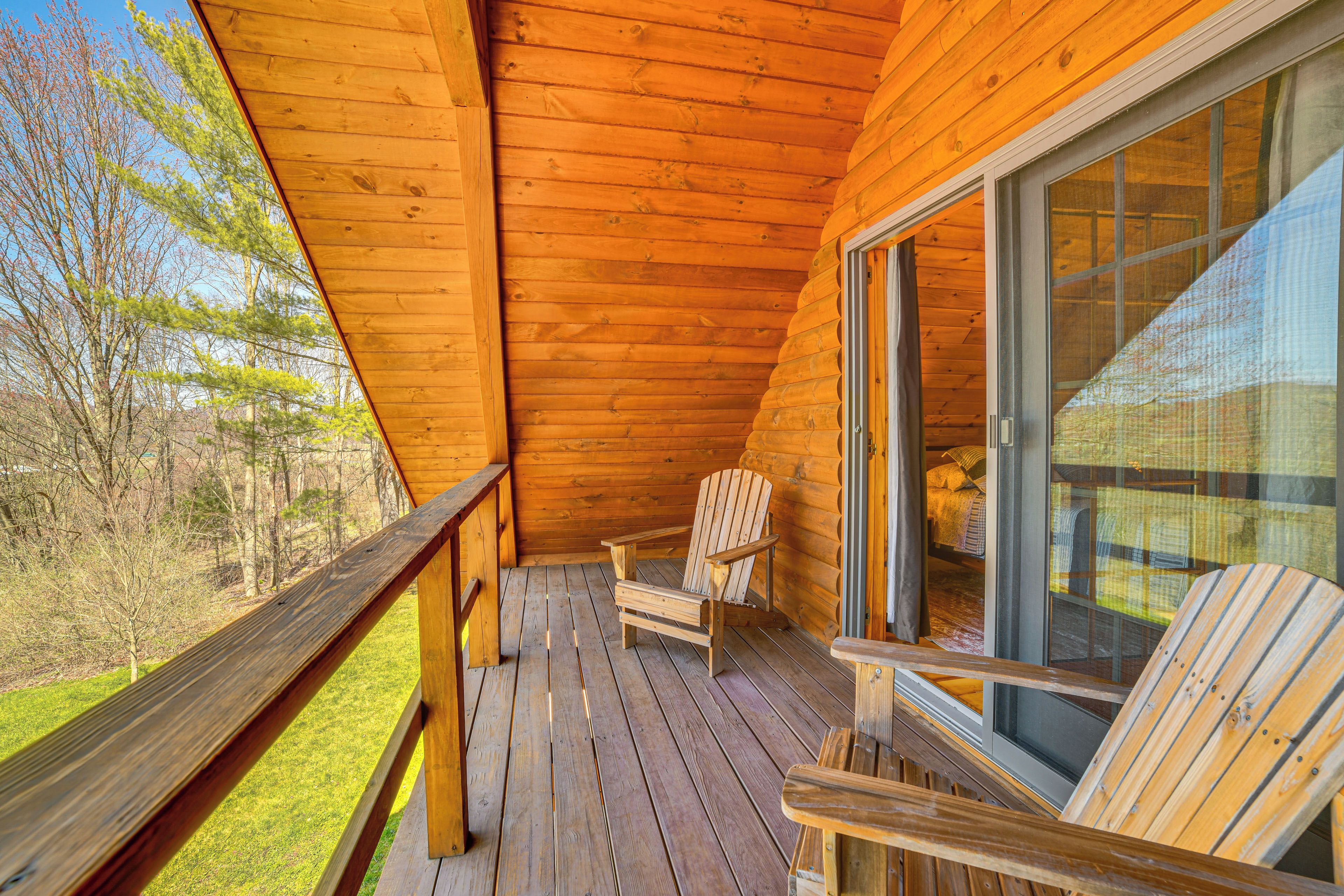 Une terrasse en bois avec deux fauteuils Adirondack, une porte vitrée et une vue sur une zone herbeuse bordée d’arbres.
