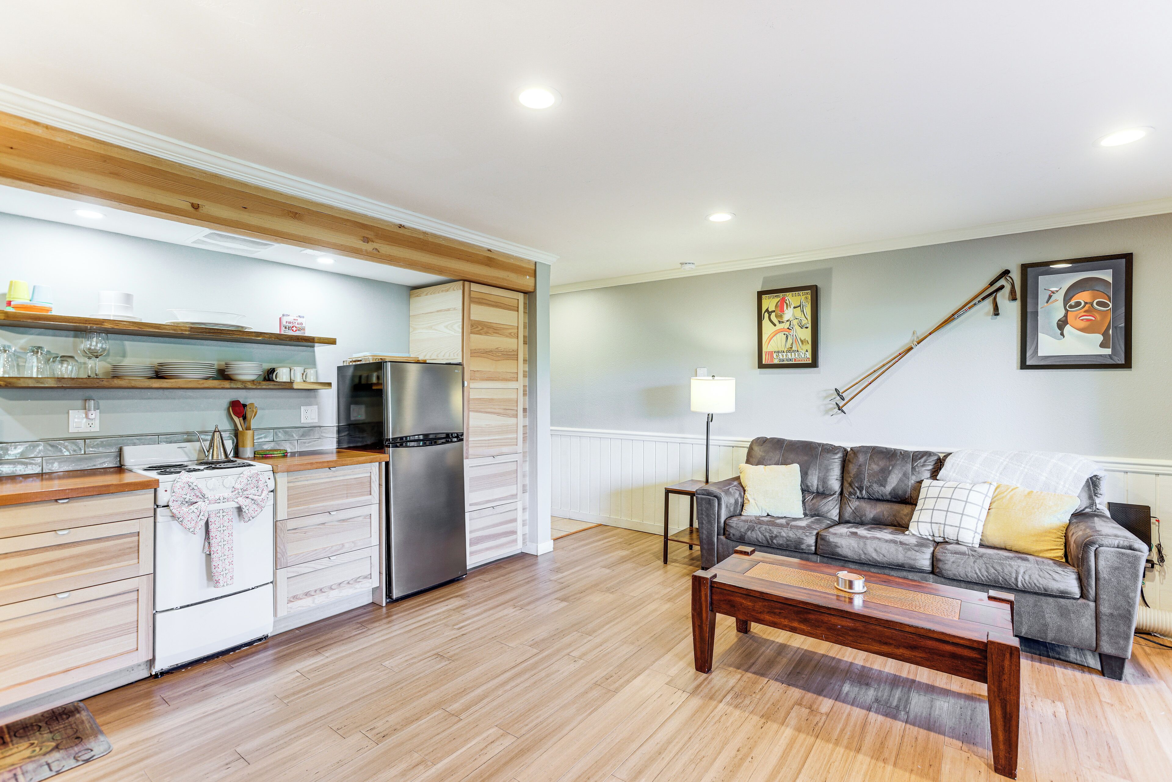 A modern kitchen with wooden cabinets, a stainless steel refrigerator, and a white stove. There is a gray sofa with cushions and a wooden coffee table in the living area.