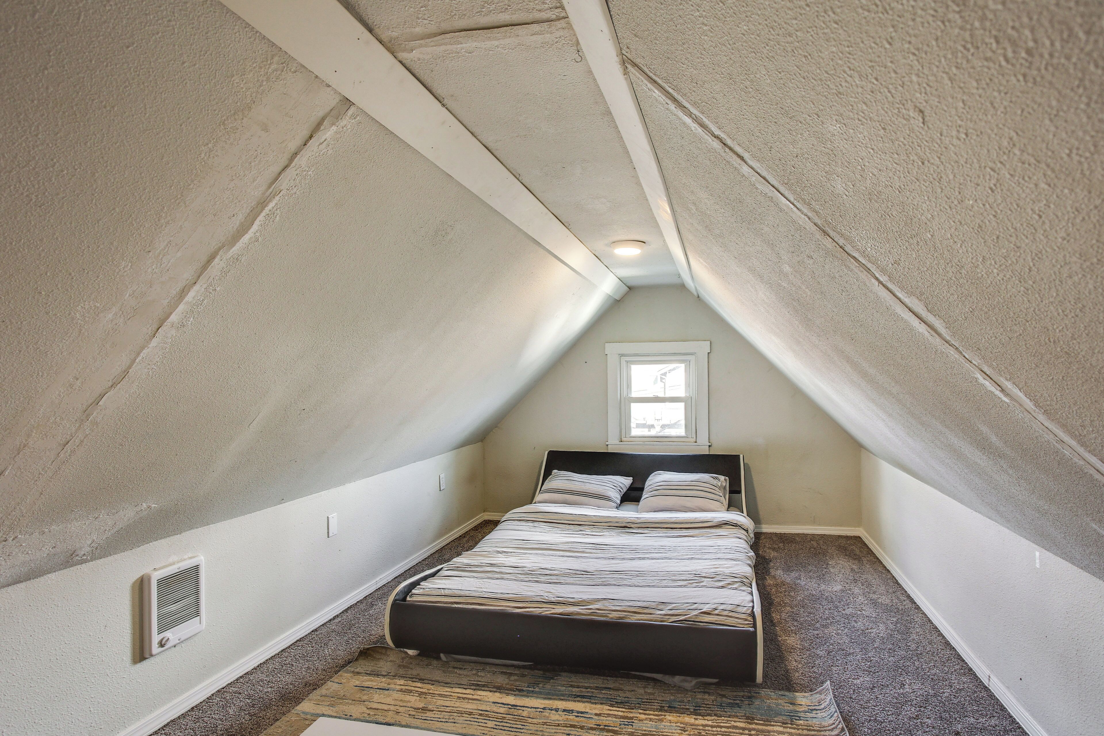 Une petite chambre mansardée de forme triangulaire, avec un lit simple, une fenêtre et une bouche d’aération au plafond.