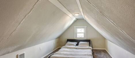 Een kleine, driehoekige slaapkamer op zolder met een enkel bed, een raam en een ventilatie rooster.