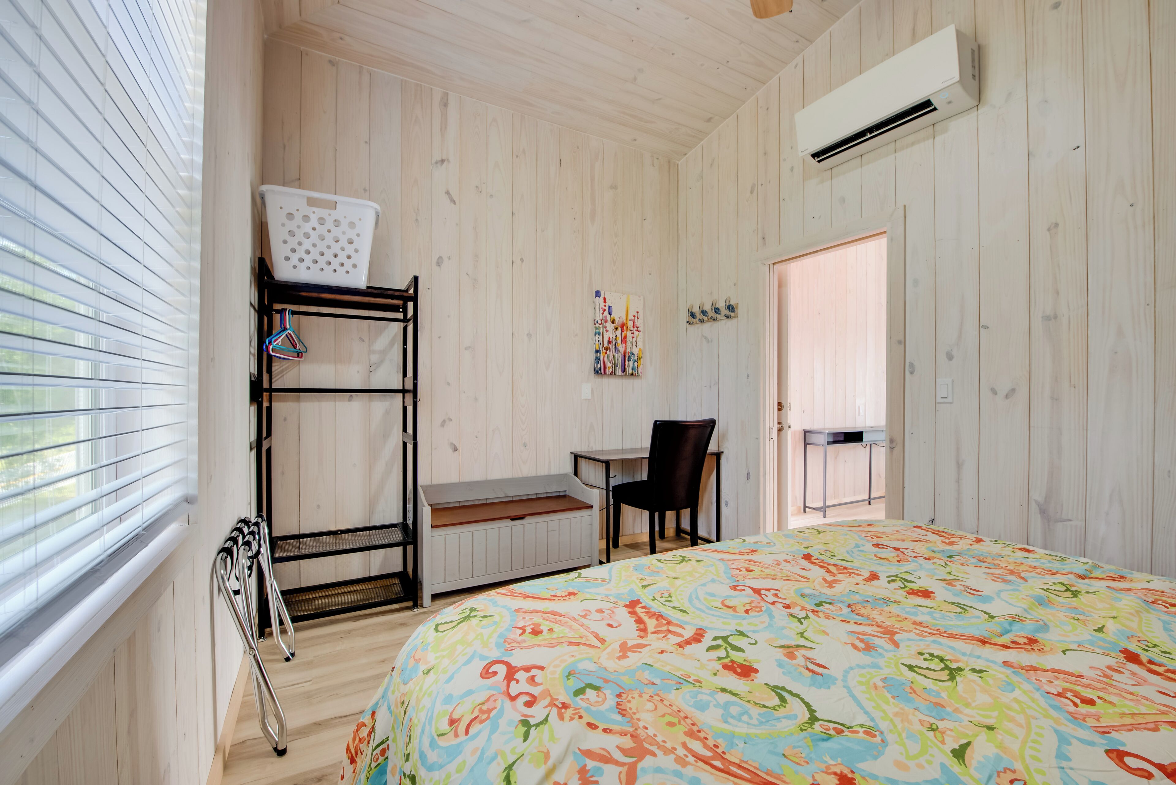 Une chambre comprenant un lit, un bureau, une chaise, une fenêtre avec des stores, un panier à linge et un appareil de climatisation.