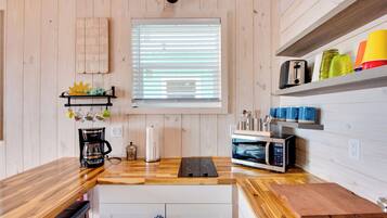 A modern kitchen with wooden walls, a window with blinds, a coffee maker, a microwave, and a shelf with colorful cups.