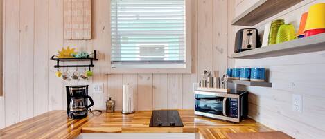 A modern kitchen with wooden walls, a window with blinds, a coffee maker, a microwave, and a shelf with colorful cups.