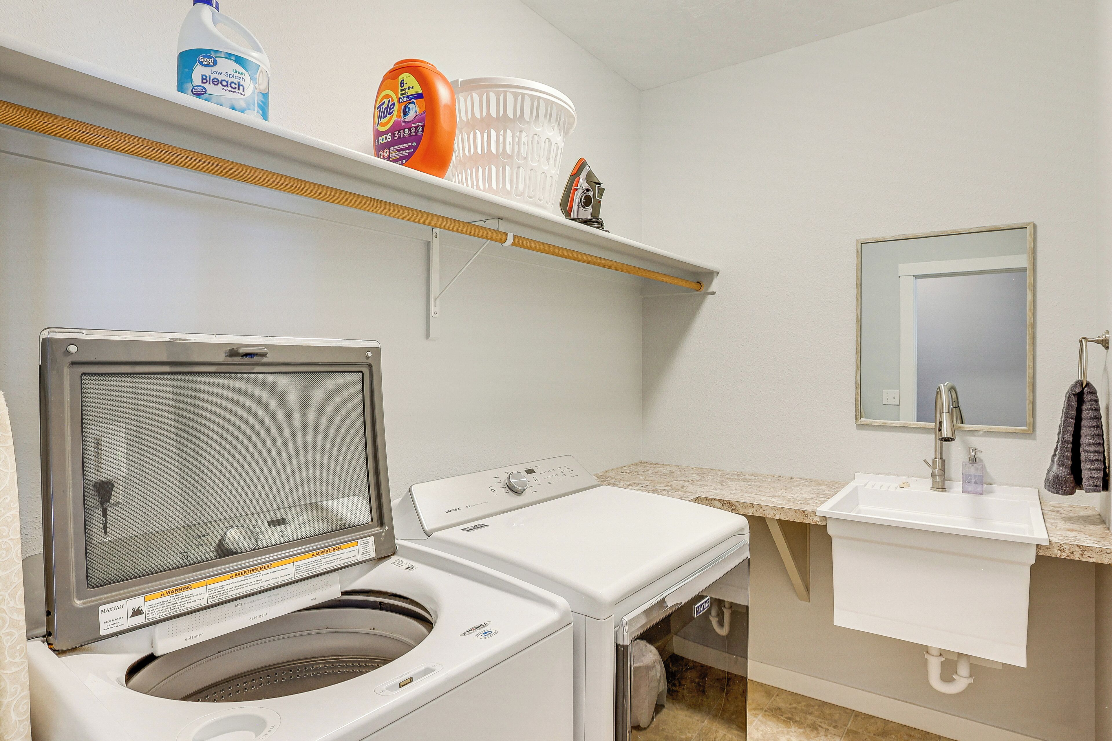 Un espace buanderie comprenant un lave-linge et un sèche-linge à chargement frontal, une étagère avec des produits de nettoyage, un évier avec un miroir et un porte-serviettes.