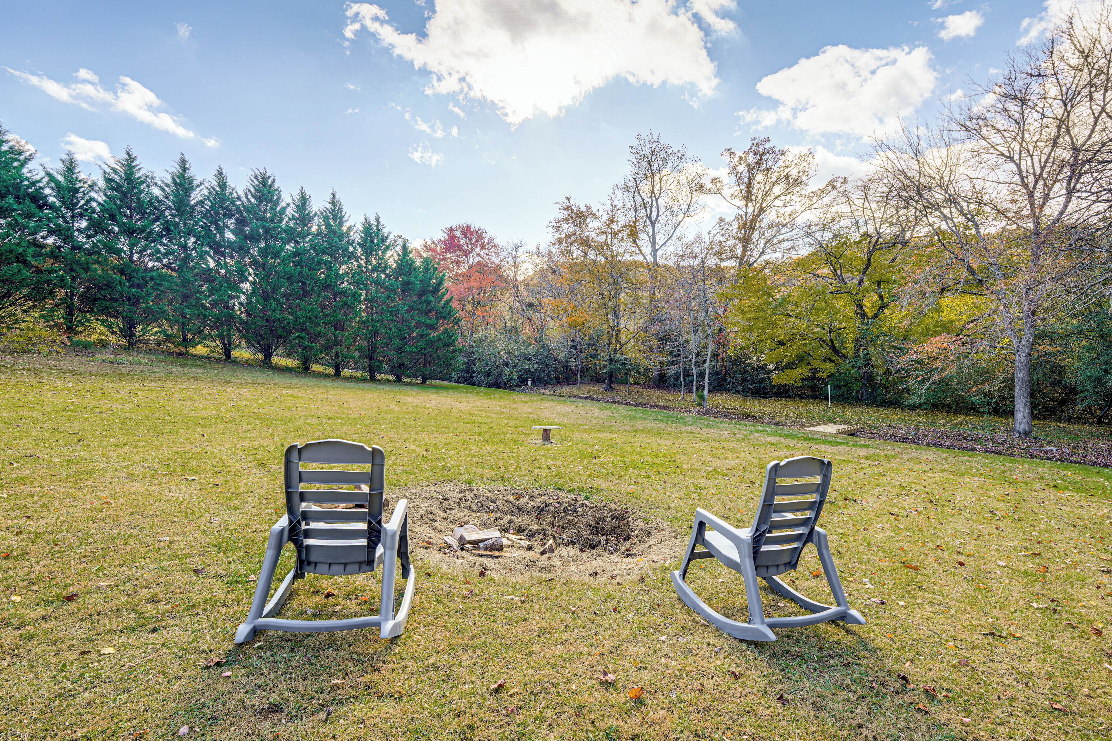 An outdoor seating area with two rocking chairs facing a fire pit surrounded by a grassy field and trees.