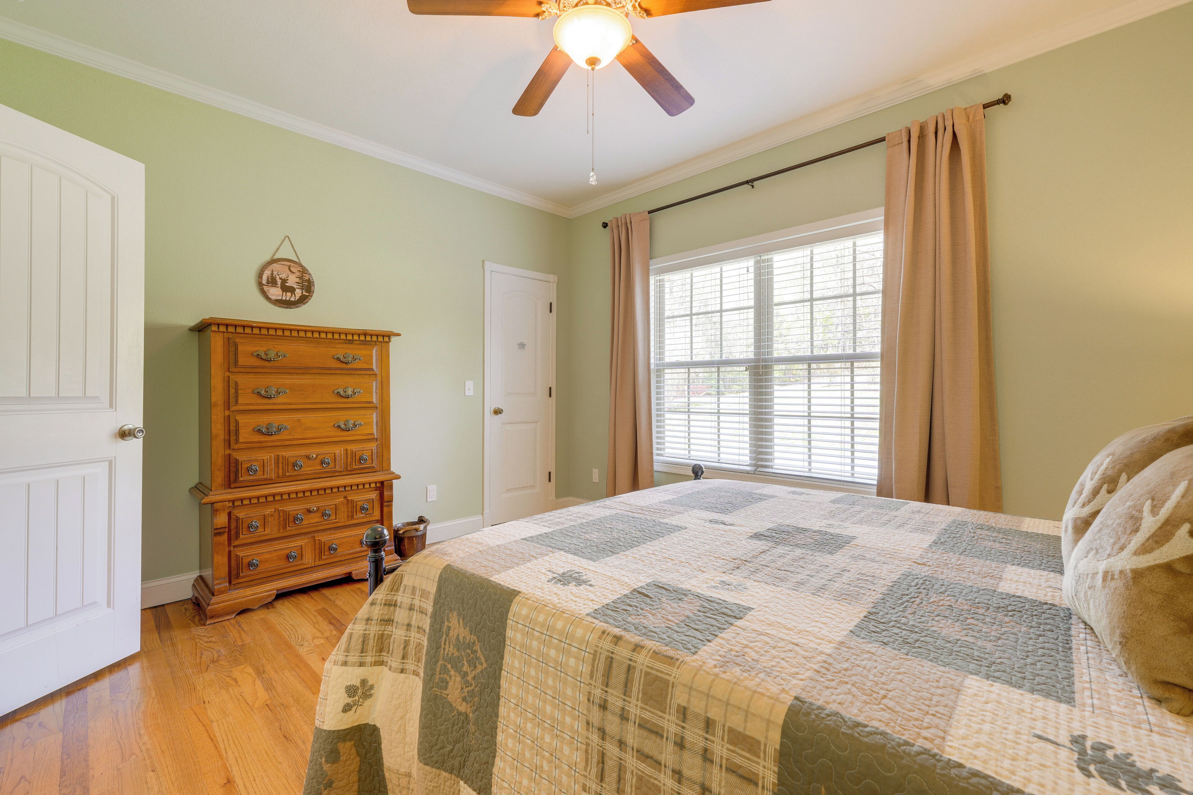 A bedroom with a wooden dresser, a quilted bedspread, a ceiling fan, and a window with curtains.