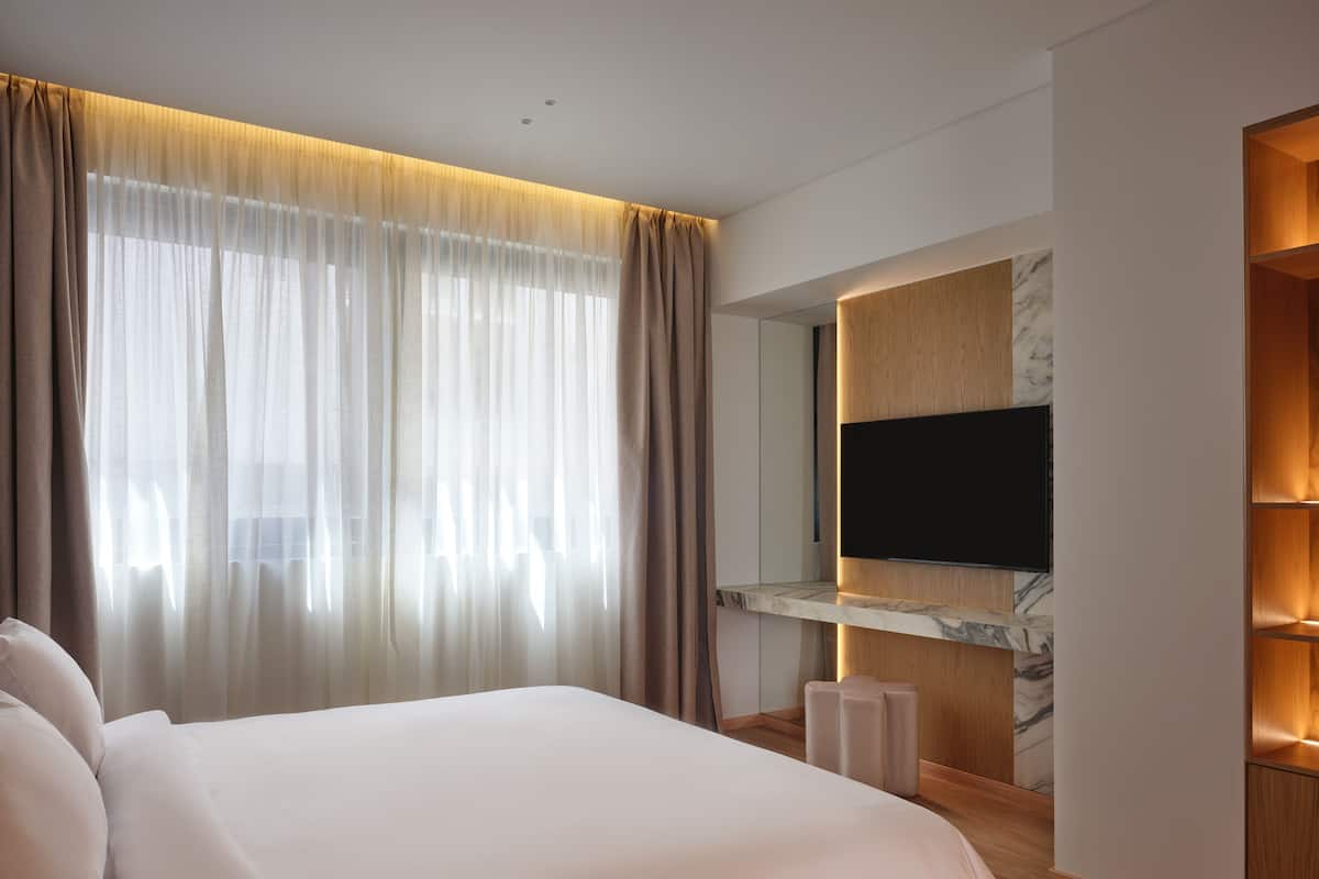 Ein modernes Hotelzimmer mit einem großen Bett, einem Flachbildfernseher, der an einer Holzvertäfelung angebracht ist, und einem großen Fenster mit transparenten Vorhängen.