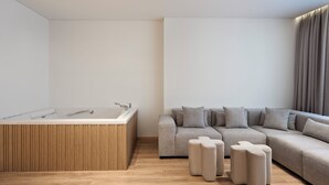 A modern bathroom with a freestanding bathtub, a wooden vanity, and a beige sofa with matching ottomans. - Nur City Suites (Athens)