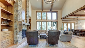 A spacious living room with a stone fireplace, wooden beams, and large windows offering a view of the outdoors. - Caldera Luxury Retreat with Access to Forest House (Sunriver)