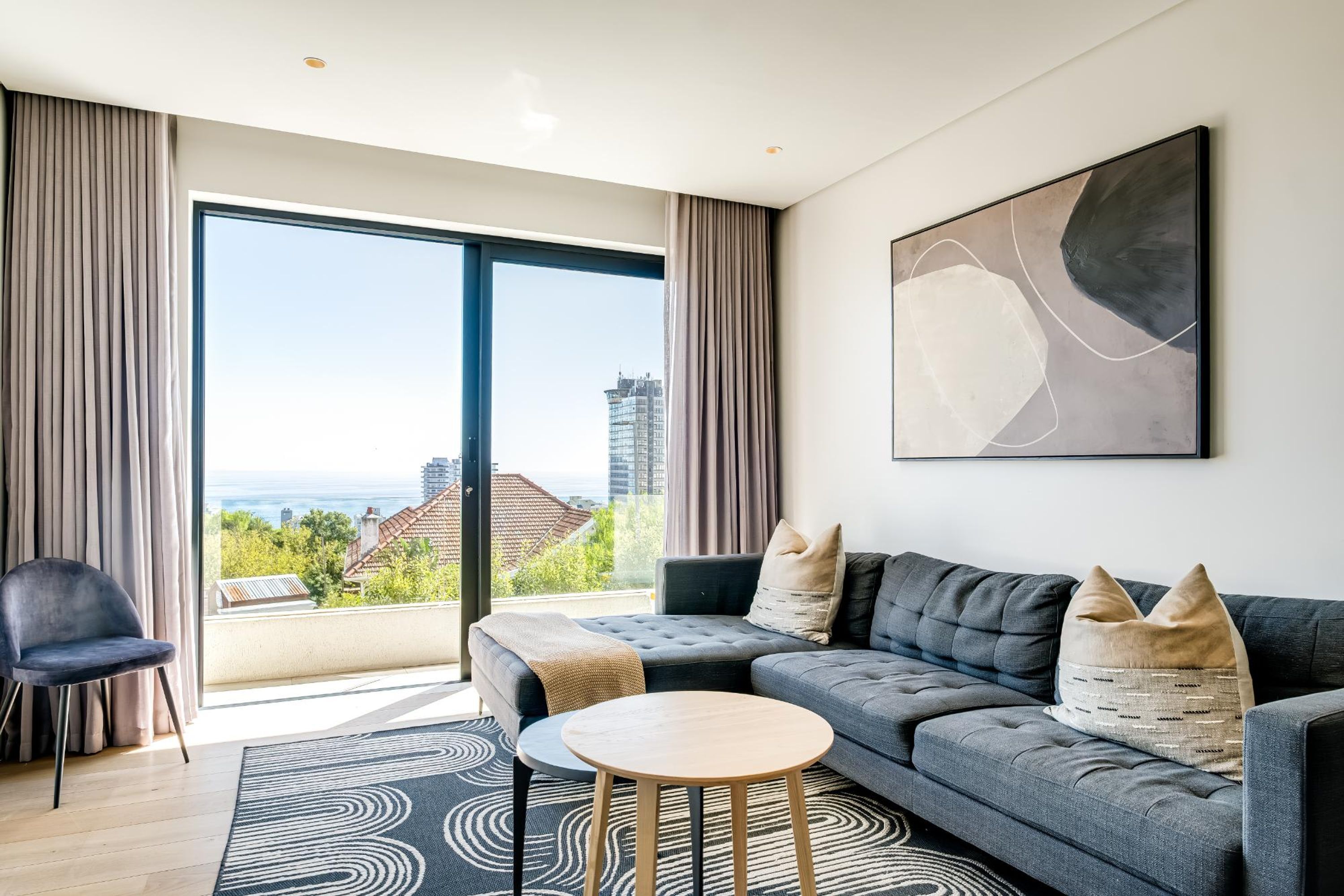 A modern living room with a grey sofa, a round wooden table, and a view of a cityscape through large windows.