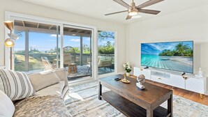 A spacious living room with a large TV, a ceiling fan, and a view of a tropical beach through large sliding doors. - Spacious Retreat With Scenic Mountain Views (Santa Ana)