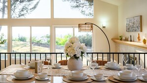 A dining table set with white plates, bowls, and glasses, a vase of white flowers, and a view of greenery through large windows. - Spacious Retreat With Scenic Mountain Views (Santa Ana)
