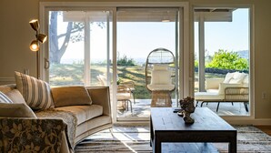 Een woonkamer met een bank, salontafel en uitzicht door grote schuifdeuren op een terras met rieten meubels en een witte stoel.