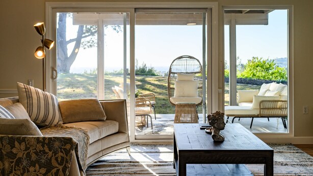 A living room with a sofa, coffee table, and a view through large sliding glass doors to an outdoor patio area with wicker furniture and a white chair. - Spacious Retreat With Scenic Mountain Views (Santa Ana)