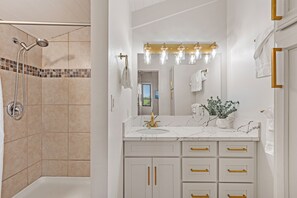 Een badkamer met een douche, een wastafel met gouden handgrepen, een spiegel met verlichting en een potplant.