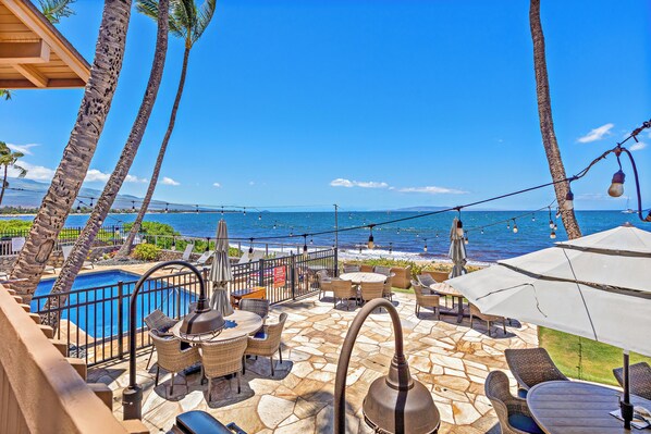 A poolside area with outdoor seating, a view of the ocean, and palm trees.