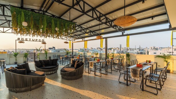 An outdoor seating area with wicker furniture and tables, a view of the city, and a hashtag sign. - The Hosteller Bangalore, Marathahalli (Bengaluru)