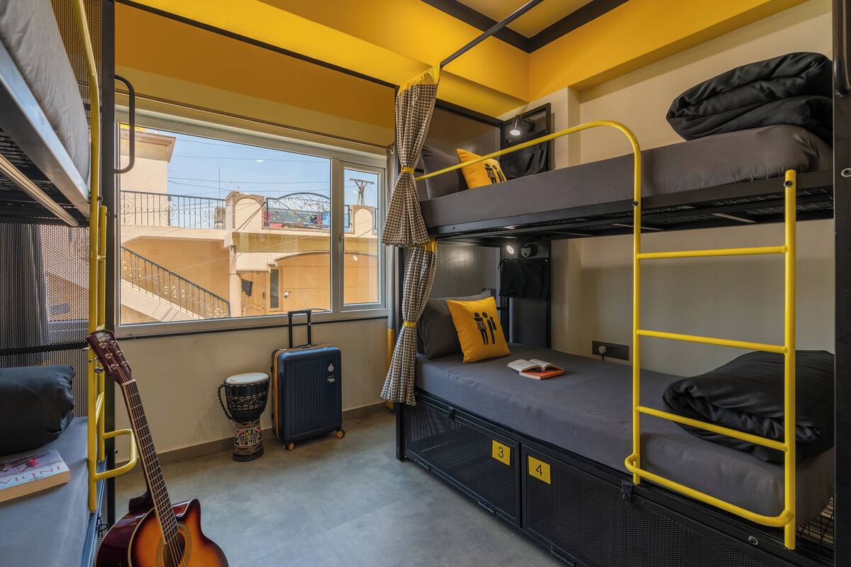 Ein kleines Zimmer mit Etagenbetten, einer Gitarre, einem Koffer und Blick auf Gebäude.