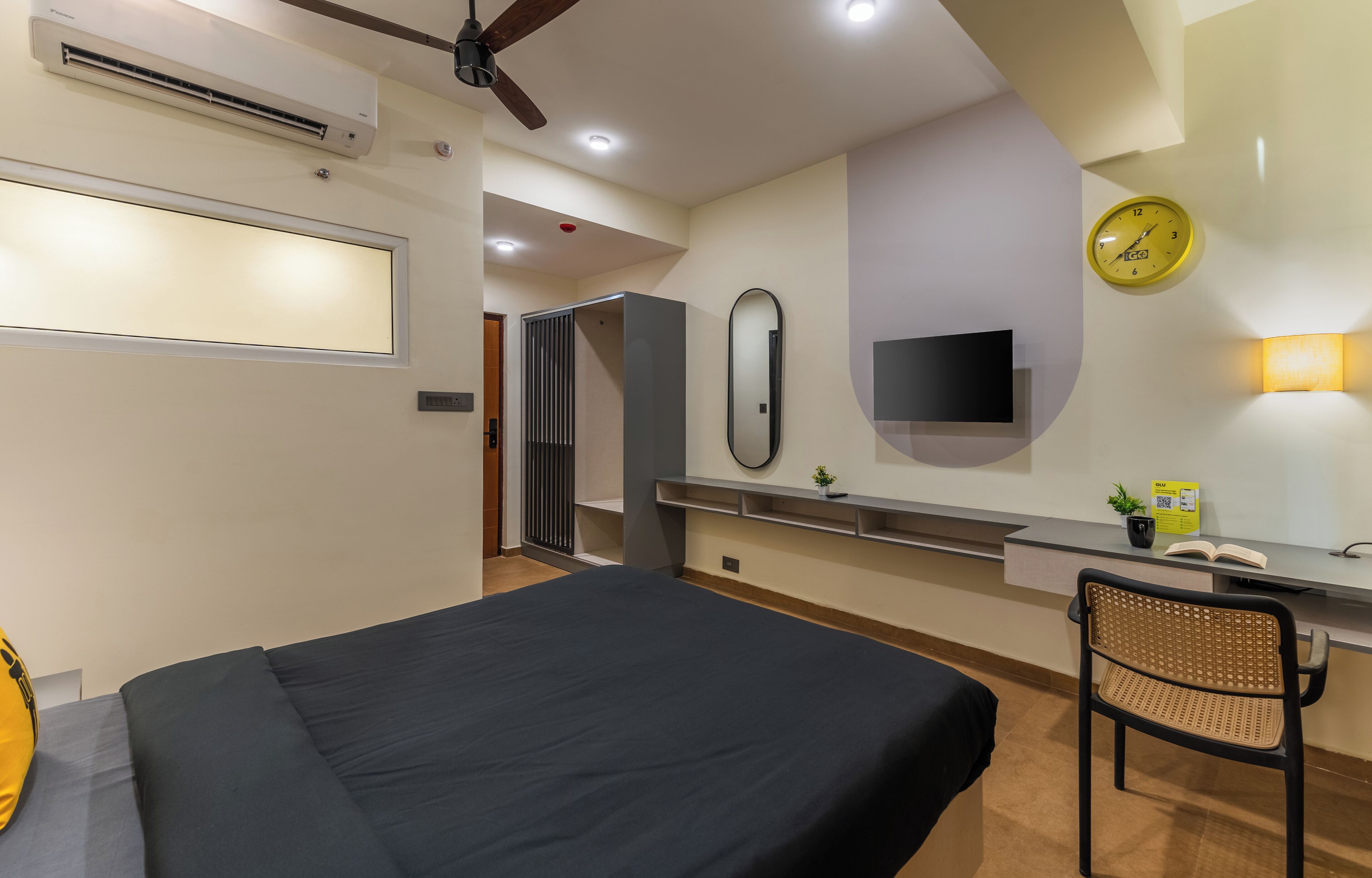 A modern hotel room with a bed, a desk with a chair, a wall-mounted TV, a clock, and an air conditioning unit.