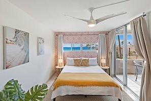 A hotel room with a bed, a ceiling fan, a view of the ocean, and a balcony with chairs.