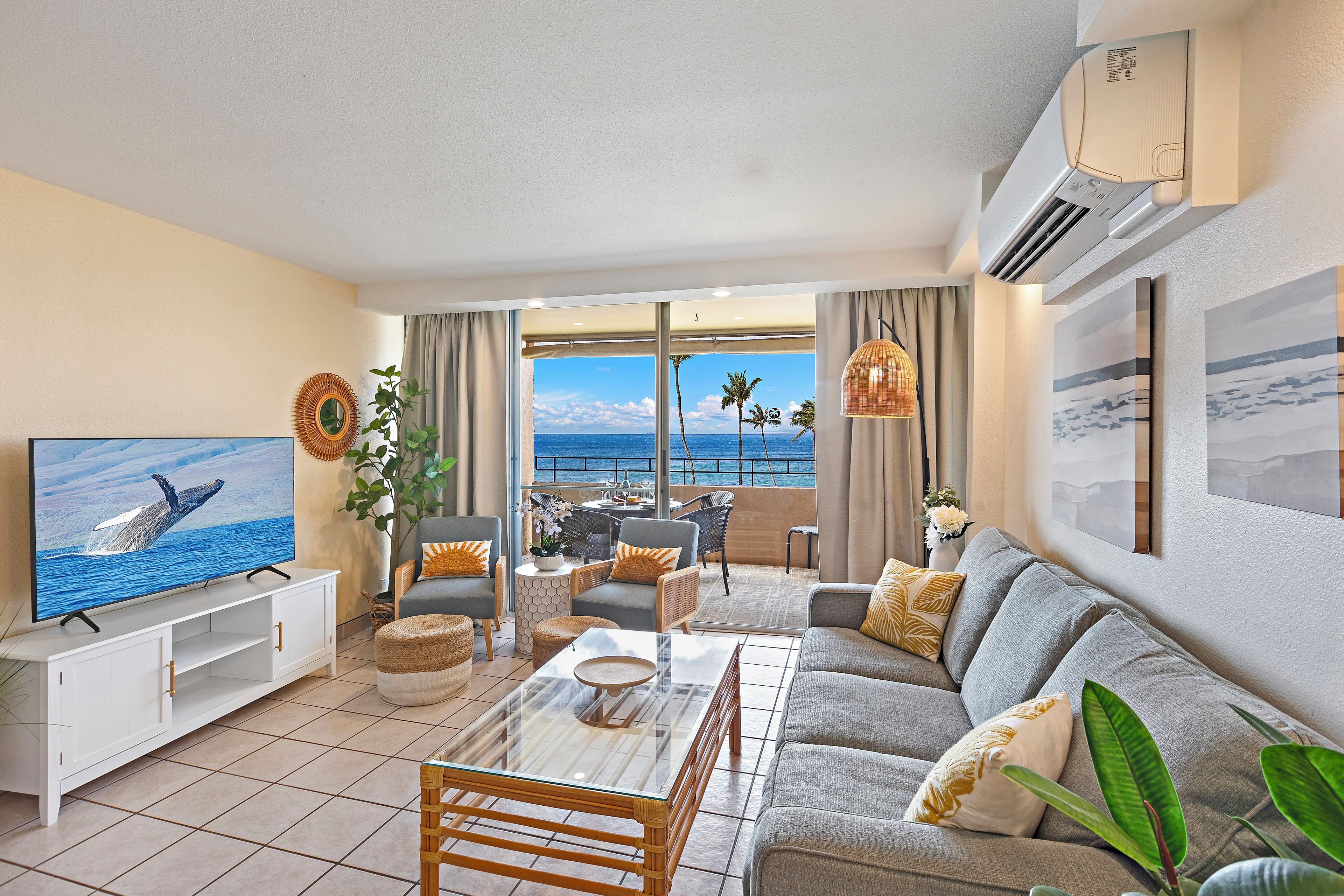 A living room with a large TV, comfortable seating, a balcony with a view of the ocean, and an air conditioning unit.