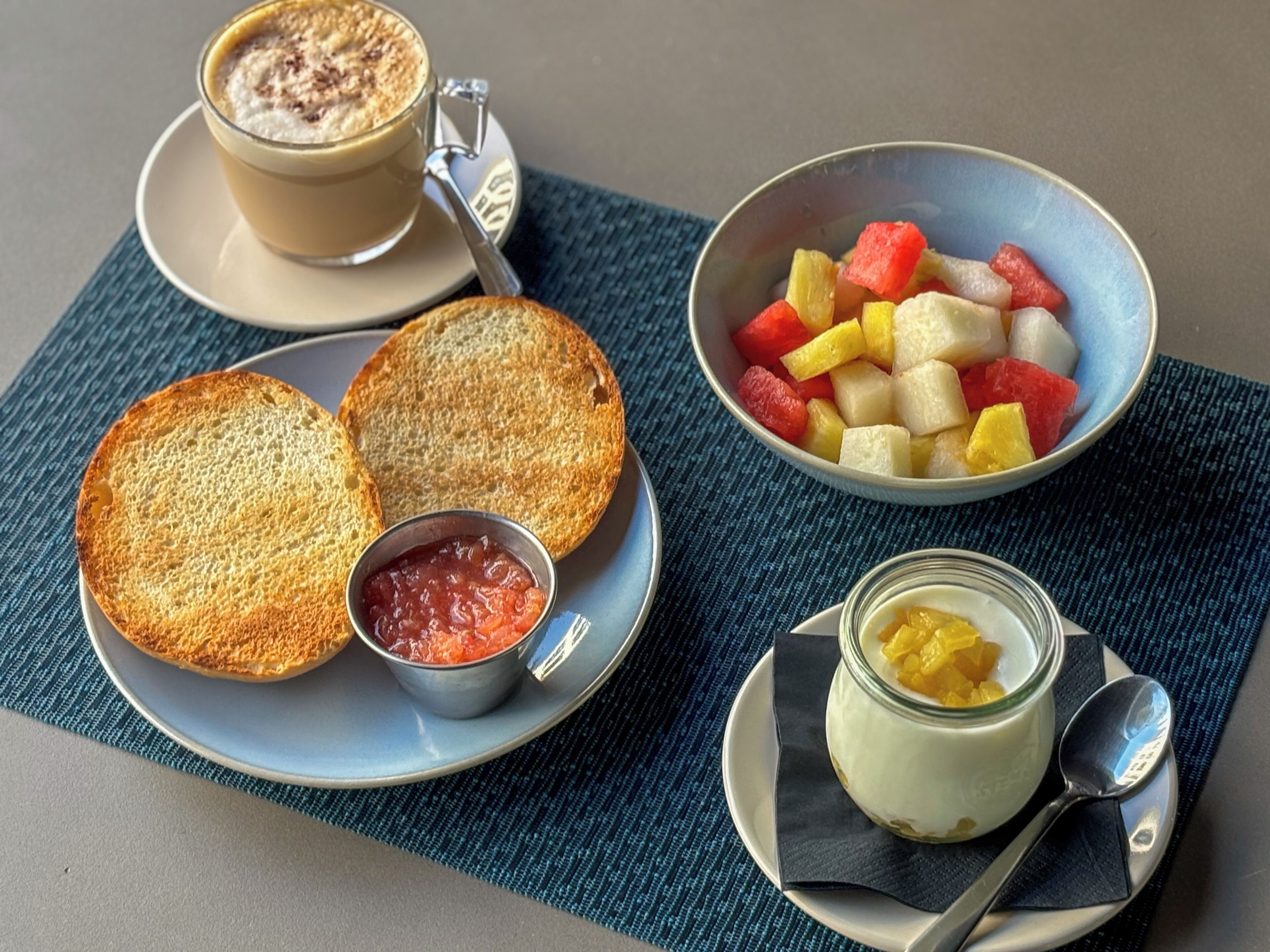 A breakfast setting with toast, jam, a cup of coffee, a bowl of fruit, and a yogurt dish.