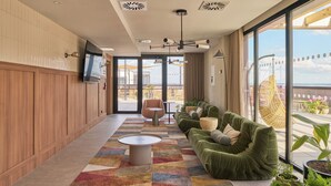 A modern hotel lobby with a wooden panel wall, a large TV, a green sofa, a patterned rug, and large windows offering a view of the outdoors. - The Carabanchel Episode (Madrid)