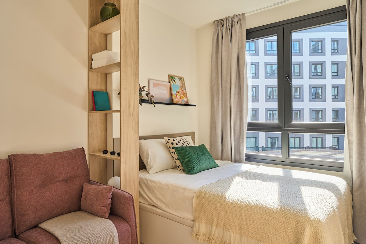 Un dormitorio con cama, sofá, ventana con cortinas y vistas a edificios.