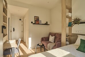 A modern, well-lit room with a wooden floor, a sofa, a bed, a small table, and a chair. There are shelves with decorative items and a painting on the wall. - The Carabanchel Episode (Madrid)