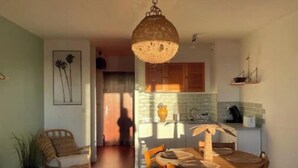 A dining area with a wooden table and chairs, a hanging light fixture, and a kitchen in the background. - Large studio with sea view - Terrace & private parking (Saint-Cyr-sur-Mer)