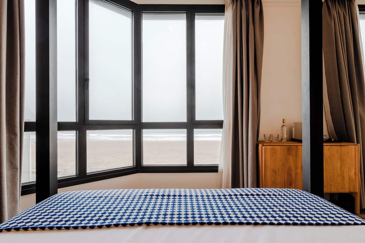 Ein Hotelzimmer mit großem Fenster und Blick auf den Strand, ein Nachttisch aus Holz mit Flasche und Gläsern sowie eine Bettdecke mit Karomuster.