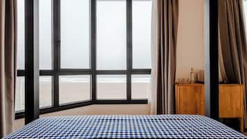 Habitación de hotel con un ventanal grande que ofrece vistas a la playa, una mesita de noche de madera con una botella y vasos, y una colcha de cuadros.