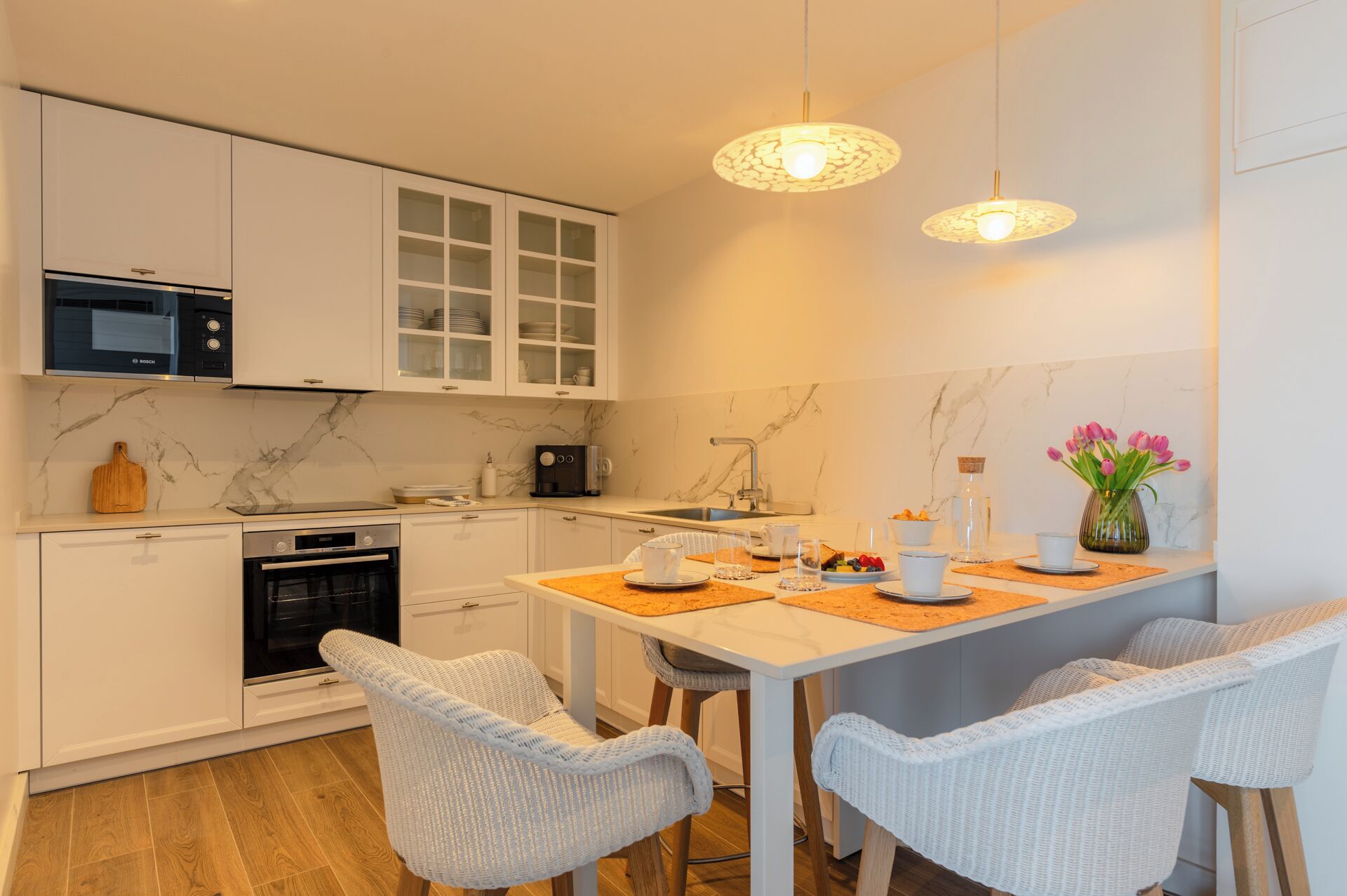 A modern kitchen with white cabinets, a Bosch microwave, and a coffee machine. There are two wicker chairs around a table with a vase of flowers.