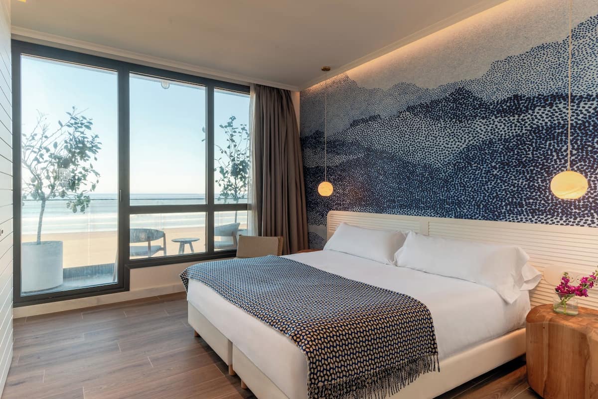 Ein Schlafzimmer mit großem Fenster mit Blick auf einen Strand, ein Bett mit gemustertem Überwurf und eine Wand mit strukturierter Gestaltung.