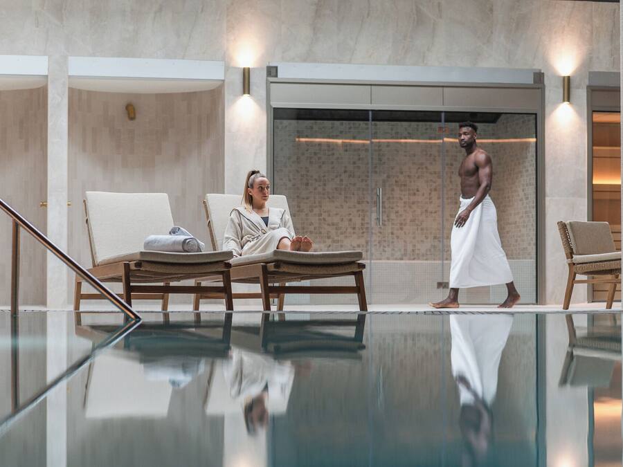 A modern indoor pool area with lounge chairs and a glass barrier.