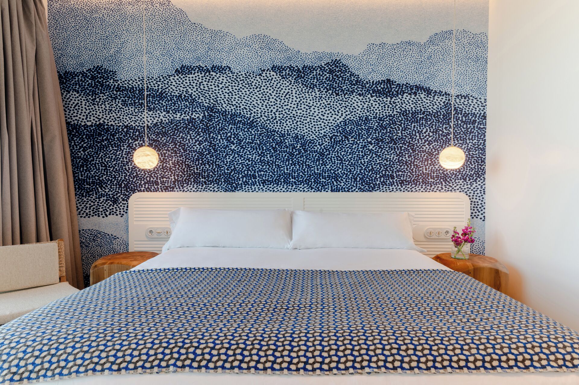A hotel room with a bed, a headboard, a wall with a dot pattern, and two hanging lights.