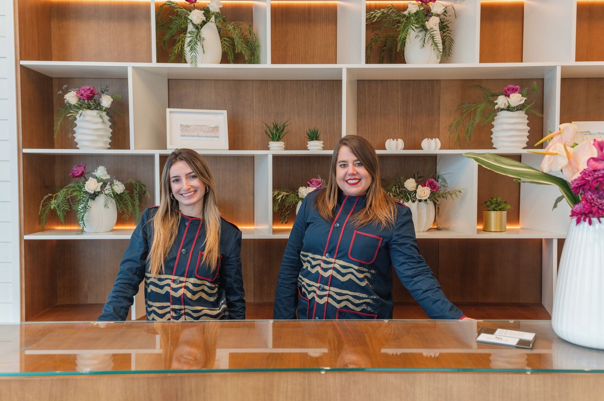 Deux réceptionnistes se tiennent derrière le comptoir d’accueil d’un hôtel, qui est agrémenté de panneaux et d’étagères en bois, sur lesquelles sont disposés des vases de fleurs et de petites plantes en pot.