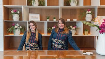 Two receptionists at a hotel reception desk with wooden paneling and shelves containing vases of flowers and small potted plants.