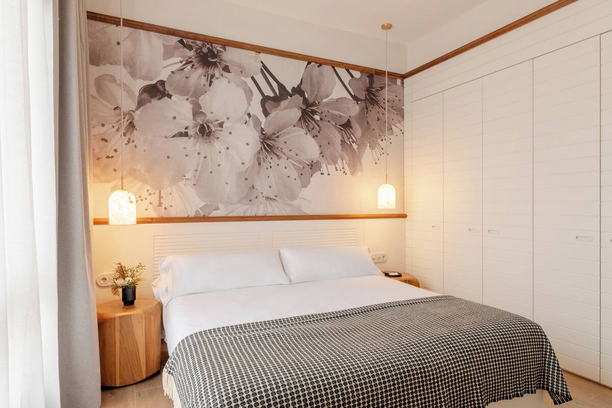 Ein Schlafzimmer mit einem großen floralen Kunstwerk, zwei Nachttischlampen, einem hölzernen Nachttisch und einer gemusterten Bettdecke.