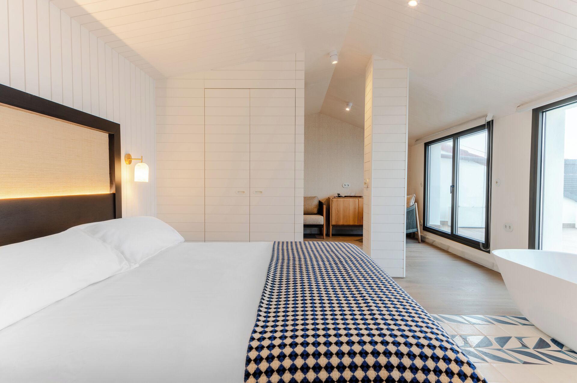 A modern hotel room with a large bed, a patterned rug, a wooden headboard, and a view through a large window.