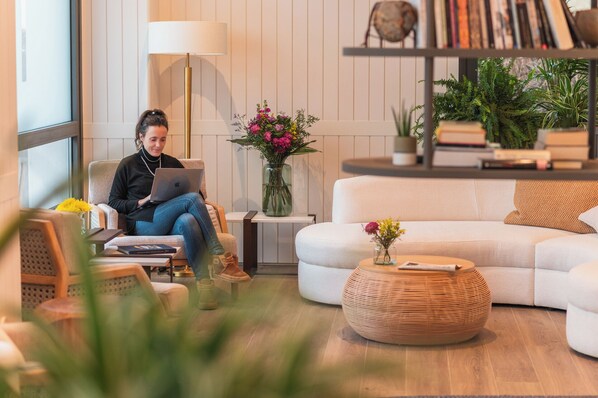 A modern living room with a person using a laptop, a white sofa, a round wicker coffee table, and a bookshelf with books and plants. - HIRU UHINAK HOTEL  (Deba)