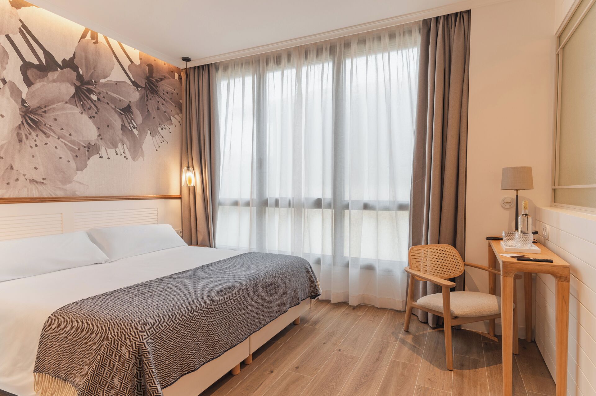 Una habitación de hotel con cama, escritorio, silla y un amplio ventanal con cortinas.