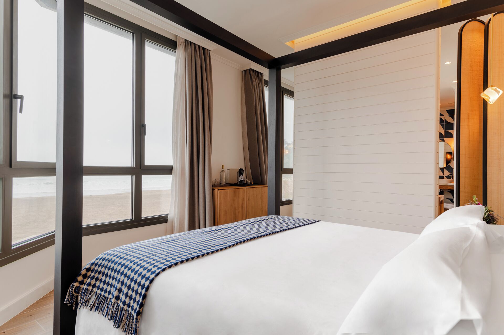 Ett hotellrum med en stor säng, utsikt över stranden och ett träskåp med en flaska och några glas på.