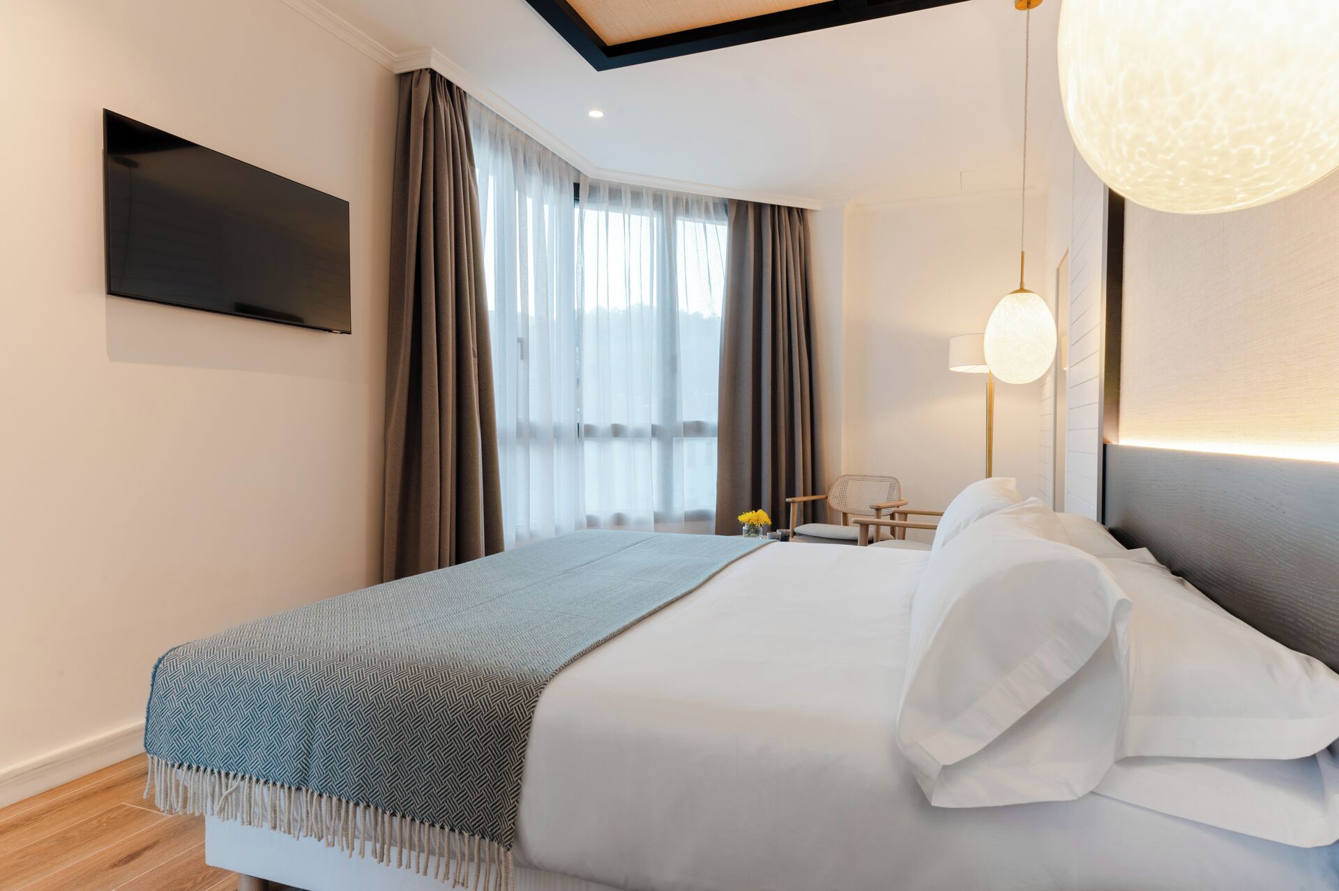 Una habitación de hotel moderna con una cama grande, un televisor de pantalla plana montado en la pared, una mesita de noche con lámpara y vistas al exterior a través de cortinas translúcidas.